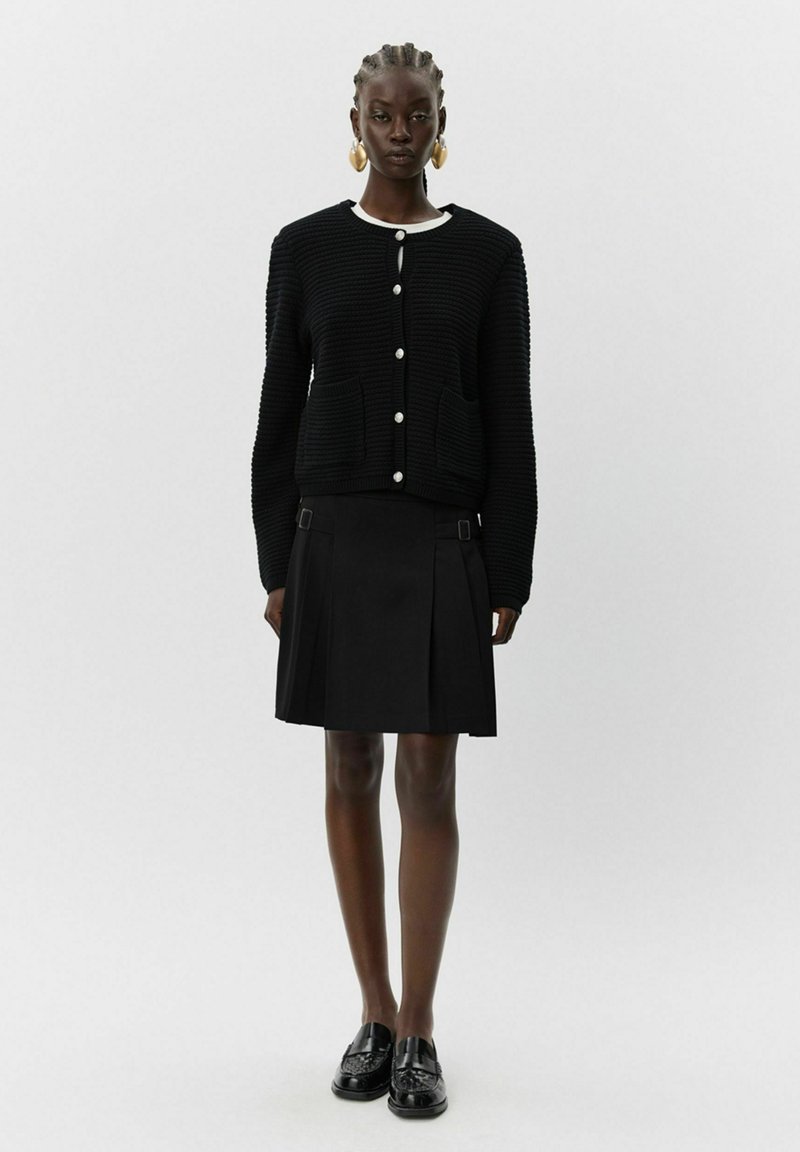 Črna pletena jakna z belim ovratnikom, z velikimi gumbi in dvema žepoma, kombinirana s črno nabranih krilom in loafersi.