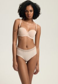 Kobieta z kręconymi włosami, nosząca cielisty stanik bez ramiączek i majtki z wysokim stanem ozdobione koronką, stojąca na jednolitym beżowym tle.