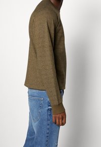 Olijfgroen, getextureerd trui met een ronde halslijn en geribbelde manchetten, gecombineerd met lichtblauwe denim jeans, weergegeven vanaf de zijkant.