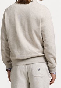 Ljusgrå sweatshirt gjord av mjukt tyg, med en ribbad krage och mudd. En liten broderad logo finns på baksidan av midjan.