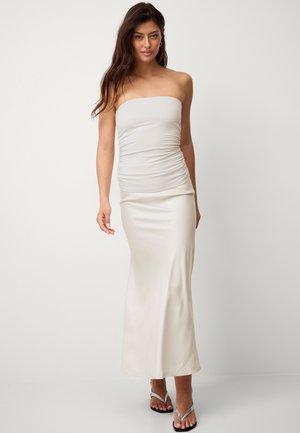 Vestido maxi sin tirantes de color beige claro, confeccionado en una tela suave y elástica, con detalles fruncidos en la cintura, que presenta una silueta ajustada y elegante.