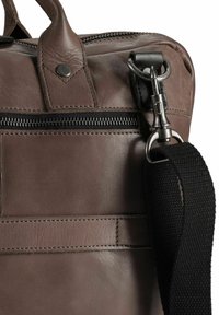 Segment van een bruine leren tas met een ritsluiting, contrasterende zwarte stiksels en een afneembare zwarte stoffen schouderband met metalen beslag.