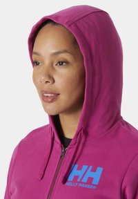 Sudadera rosa con cremallera, capucha con cordón y un logo azul "HH" en el pecho. Textura de tela suave, diseño casual.