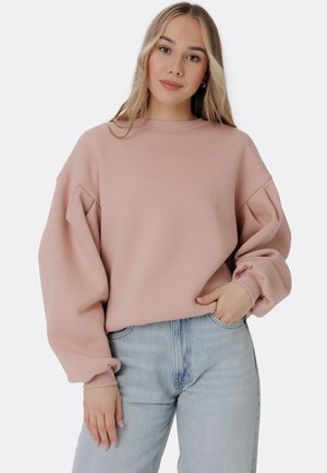 Světle růžová oversized mikina s kulatým výstřihem, měkkou texturou a spuštěnými ramenními švy, doplněná vysokými světle modrými džíny.