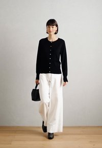 Cardigan nero a maglia con bottoni bianchi, abbinato a pantaloni bianchi larghi e calzature nere. Il modello tiene una borsa nera strutturata.