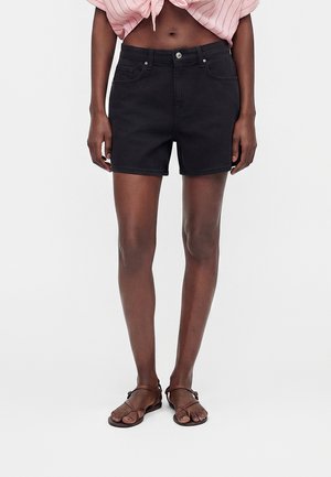 Kvinde iført sorte denimshorts, lyserød bundet crop top og brune flade sandaler med remme står på en hvid baggrund.