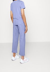 Person som står klädd i ljuslila kortärmad skjorta och matchande beskurna byxor med vita och rosa träningsskor, mot en enfärgad bakgrund.