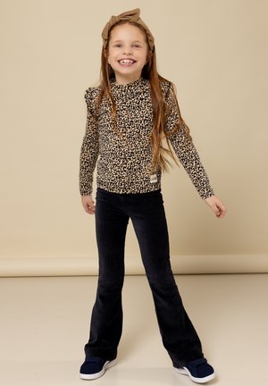 Lange mouw top met luipaardprint in bruin en zwart, gecombineerd met marineblauwe flared corduroybroek. Model draagt donkere instappers en een bruine haarband.