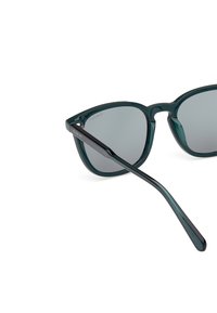 Gafas de sol de acetato verde con lentes grises y un marco angular distintivo. La varilla negra presenta un grabado de la marca. Acabado suave.
