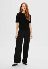 Zwarte gebreide top met korte mouwen en een ronde halslijn, gecombineerd met brede zwarte broeken en platte zwarte schoenen. Eenvoudig, gestroomlijnd ontwerp.