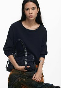 Borsa nera a spalla con texture arricciata e dettagli metallici. La modella indossa un maglione blu navy e una gonna a fantasia e texture.