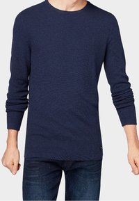Pull en tricot bleu marine avec un col rond et des manches longues, muni de poignets et d'un ourlet côtelés. Texture légère et un minimalisme au niveau de la marque.