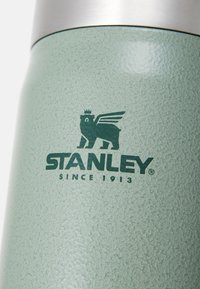 Žalias tekstūruotas nerūdijančio plieno termosas su sidabriniu dangteliu ir juodu lokio su sparnais logotipu, užrašu "STANLEY SINCE 1913."