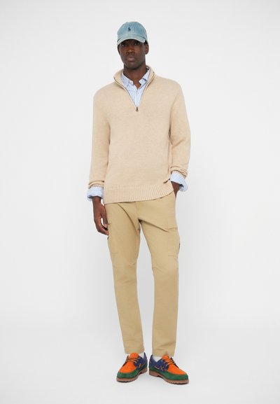 Beige kötött pulóver cipzáros gallérral, alatta csíkos kék ing, bézs cargo nadrág és színes mintás cipők. Világoskék sapka.