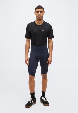 Homme portant un maillot de cyclisme noir à manches courtes avec le logo "poc", un cuissard de cyclisme bleu marine, des chaussettes noires et des baskets noires aux lacets blancs.