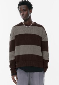 Un pull en tricot rayé marron et gris avec une texture côtelée, une coupe décontractée, un col rond et des manches longues. Assorti avec un pantalon gris.