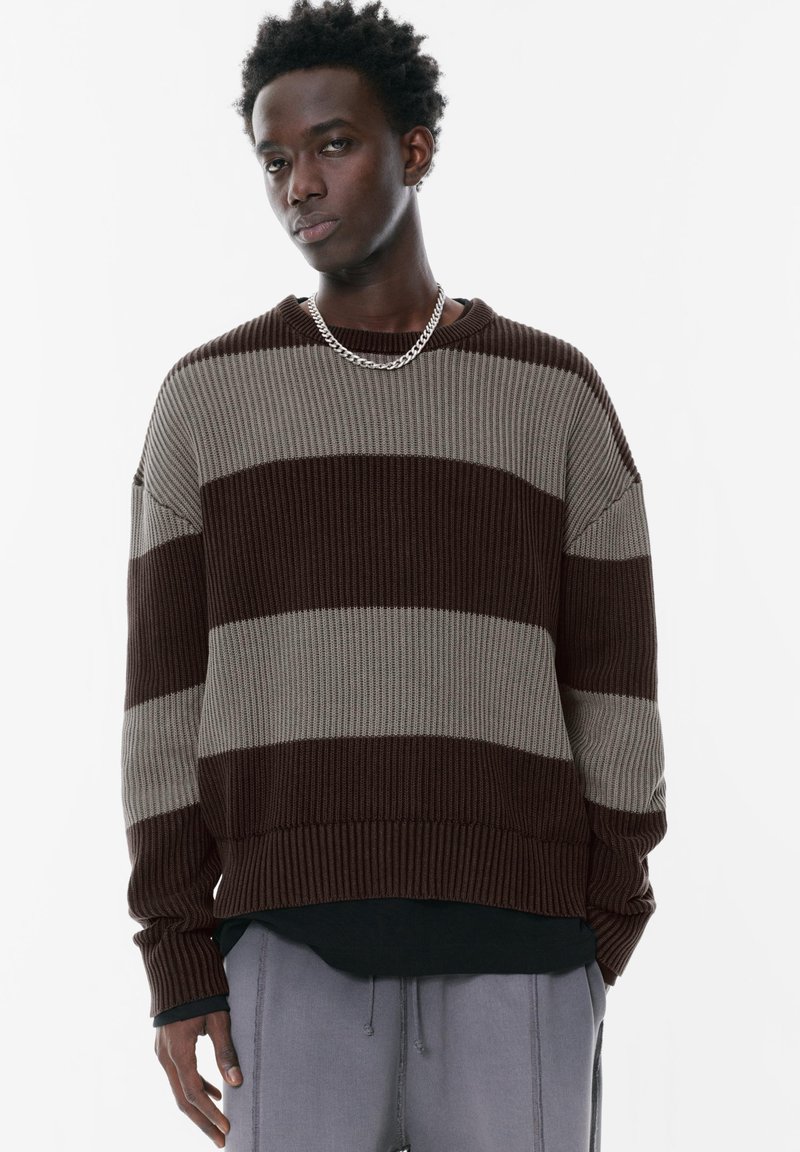 Un pull en tricot rayé marron et gris avec une texture côtelée, une coupe décontractée, un col rond et des manches longues. Assorti avec un pantalon gris.