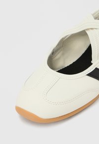 Λευκό μπαλαρίνα παπούτσι με ελαστικούς λουριάδες που διασταυρώνονται, μαύρη εσωτερική σόλα και ελαφρώς καφέ λαστιχένια σόλα, εμφανίζεται σε κοντινή λήψη σε λευκό φόντο.