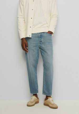 Hombre con vaqueros azul claro, zapatillas beige y una camisa crema abotonada, de pie con una mano en el bolsillo contra un fondo liso.