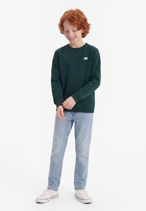 Garçon roux en sweat-shirt vert foncé, jean bleu clair et baskets blanches, debout en souriant en ajustant sa manche.