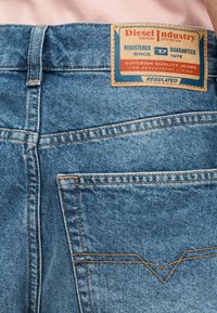 Πίσω μέρος μεσαίου μπλε τζιν με πορτοκαλί ραφές και ορθογώνια ετικέτα Diesel Industry στον ιμάντα της μέσης.