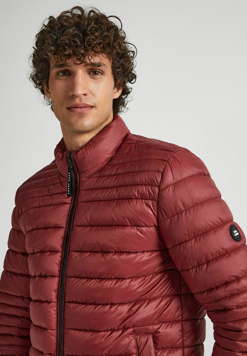 Bordowa kurtka puchowa z poziomym pikowaniem, wysoki kołnierz i czarna naszywka z logo na rękawie; gładki, lekki materiał.