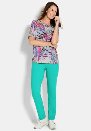 Vrouw glimlachend met hand bij gezicht, draagt een korte mouwen bloemen top, turquoise broek en witte sneakers, staand tegen een witte achtergrond.