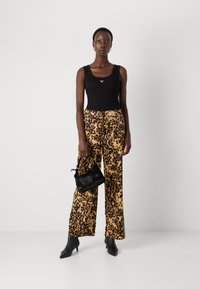 Top nero senza maniche con logo, pantaloni beige con stampa leopardata nera, tiene in mano una piccola borsa nera, e indossa tacchi neri a punta.