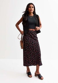 Un gilet sans manches noir sur un crop top noir ajusté, associé à une longue jupe noire avec de petits motifs floraux roses. Porté avec des sandales à talons noires.