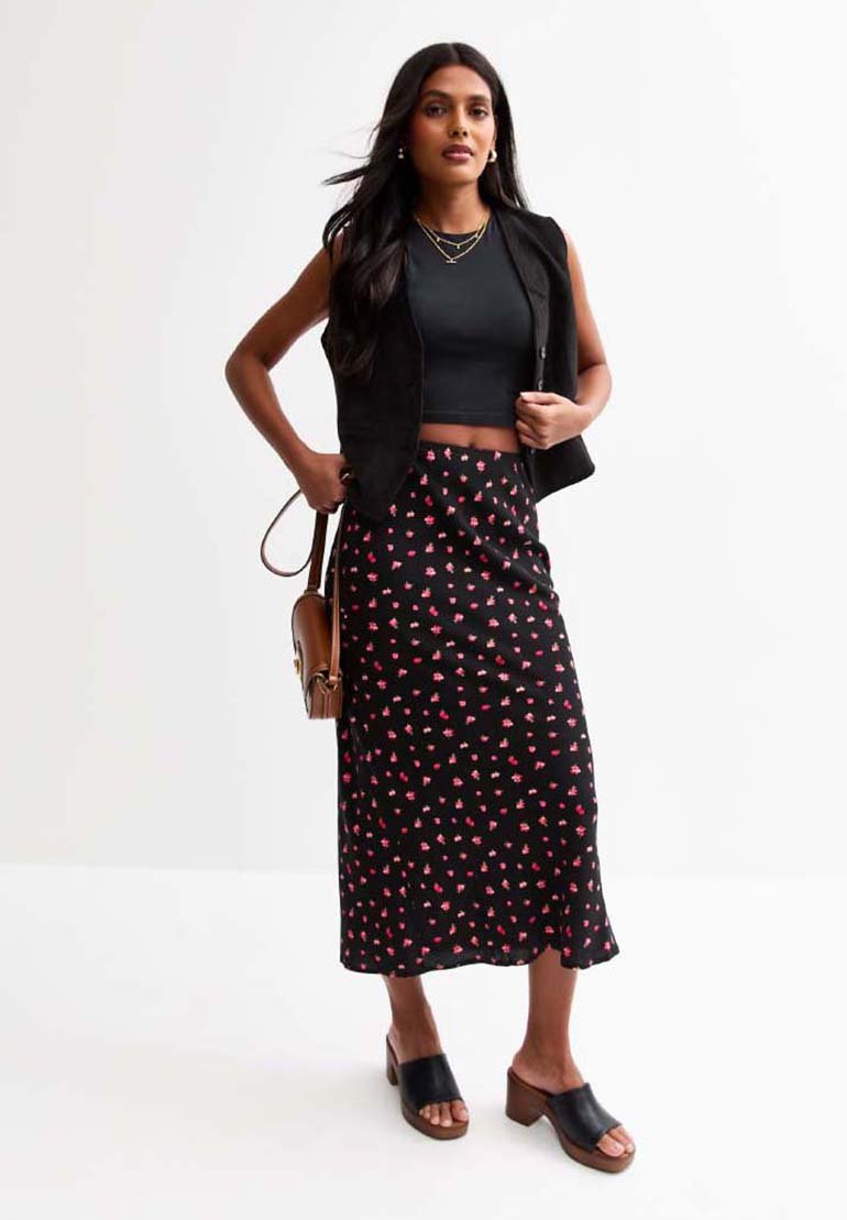 Un gilet sans manches noir sur un crop top noir ajusté, associé à une longue jupe noire avec de petits motifs floraux roses. Porté avec des sandales à talons noires.