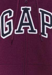La sudadera con cremallera burdeos presenta un gran logotipo de "GAP" en blanco y navy. Está hecha de una tela suave con cordones a juego y cremallera frontal.
