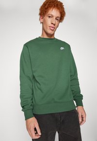 Sudadera verde de tejido suave con cuello redondo. Presenta puños y dobladillo acanalados, y un pequeño logo blanco en la parte superior izquierda del pecho.
