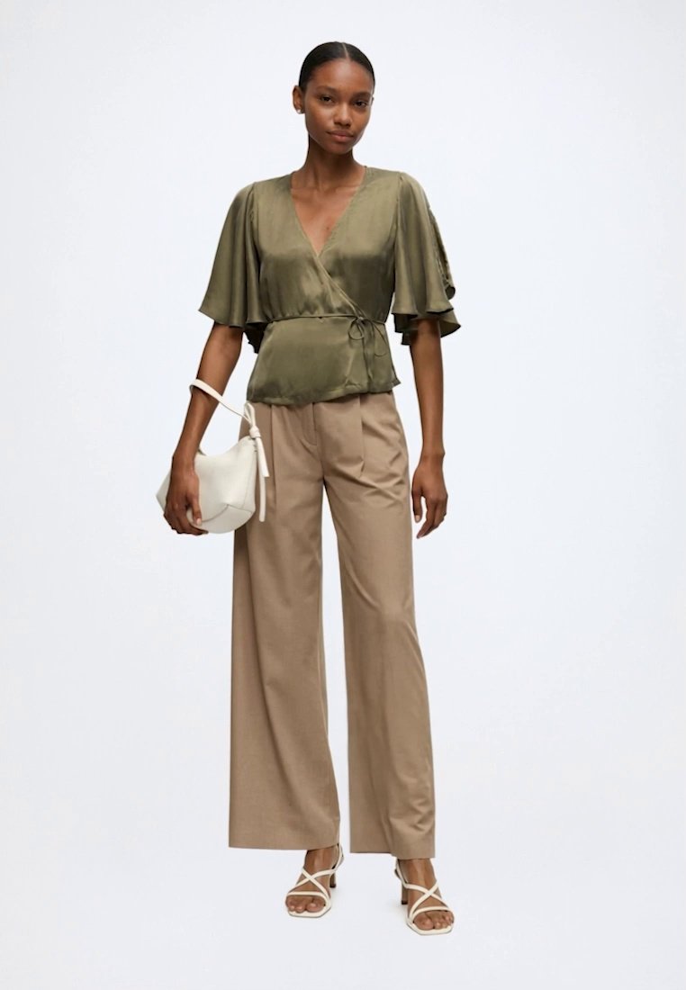 Femme portant un chemisier cache-cœur vert olive à larges manches courtes, un pantalon beige à jambes larges, des sandales blanches et tenant un sac à main blanc.