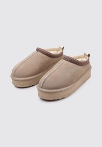 Zapatillas beige sin cordones con una textura suave, que presentan un ribete marrón con estampado, una suela acanalada y un forro acogedor para mayor comodidad.