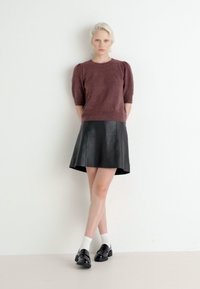 Burgundské pletené svetr s krátkým rukávem v kombinaci s černou koženou rozšířenou sukni. Modelka má na sobě bílé ponožky a černé boty, stojící před bílou zdí.