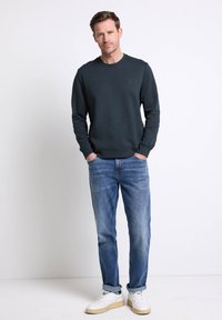 Felpa verde scuro con motivo testurizzato, collo rotondo e polsini a costine, abbinata a jeans blu chiaro e sneakers bianche con suole beige.