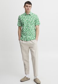 Grønn t-skjorte med korte ermer og hvitt blomstermønster, sammen med beige bukser og brune slip-on sko. Enkel krageutforming, lett stoff.