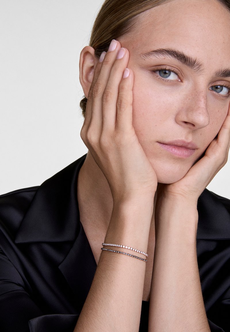 Ezüst karkötők apró, tiszta drágakövekkel, mezítelen csuklón viselve, fekete selyeming háttérrel. Simás bőrtextúra, semleges kifejezés.