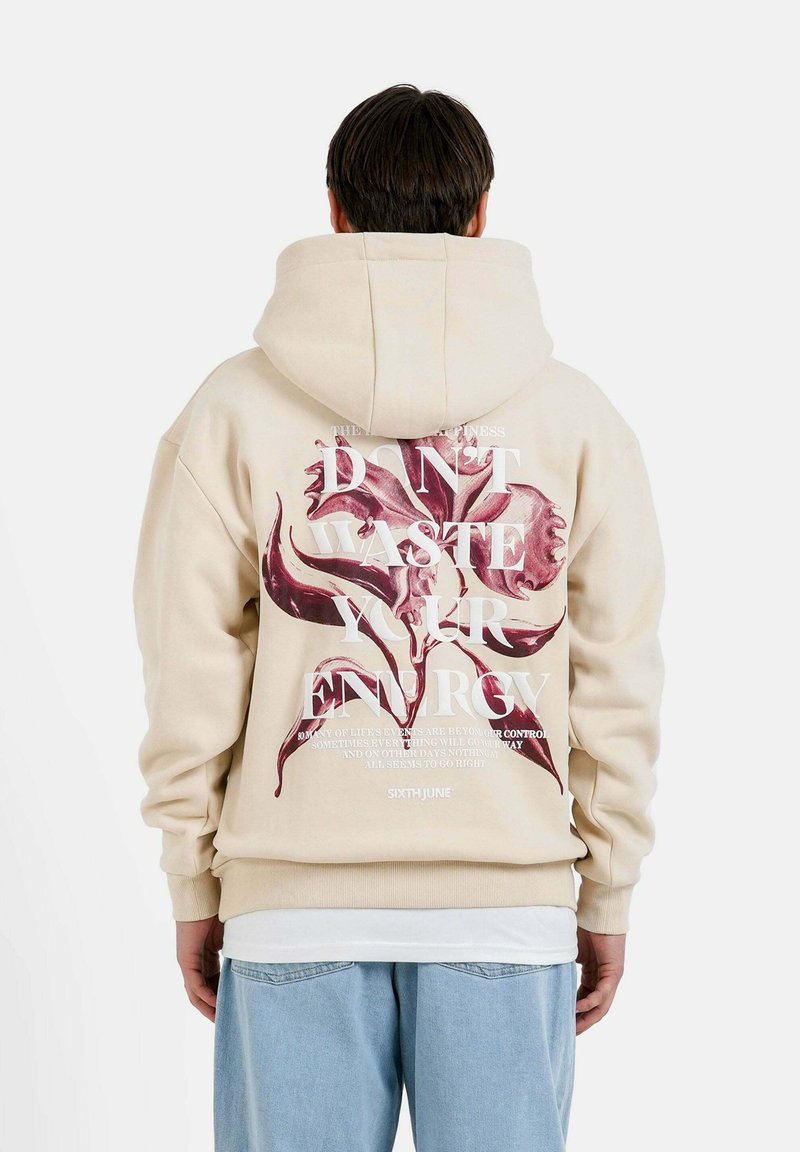 Beige hoodie med en stor blommig grafik och text på baksidan. Tillverkad i bomull, avslappnad passform, ribbade muddar och fåll, samt huva med dragsko.