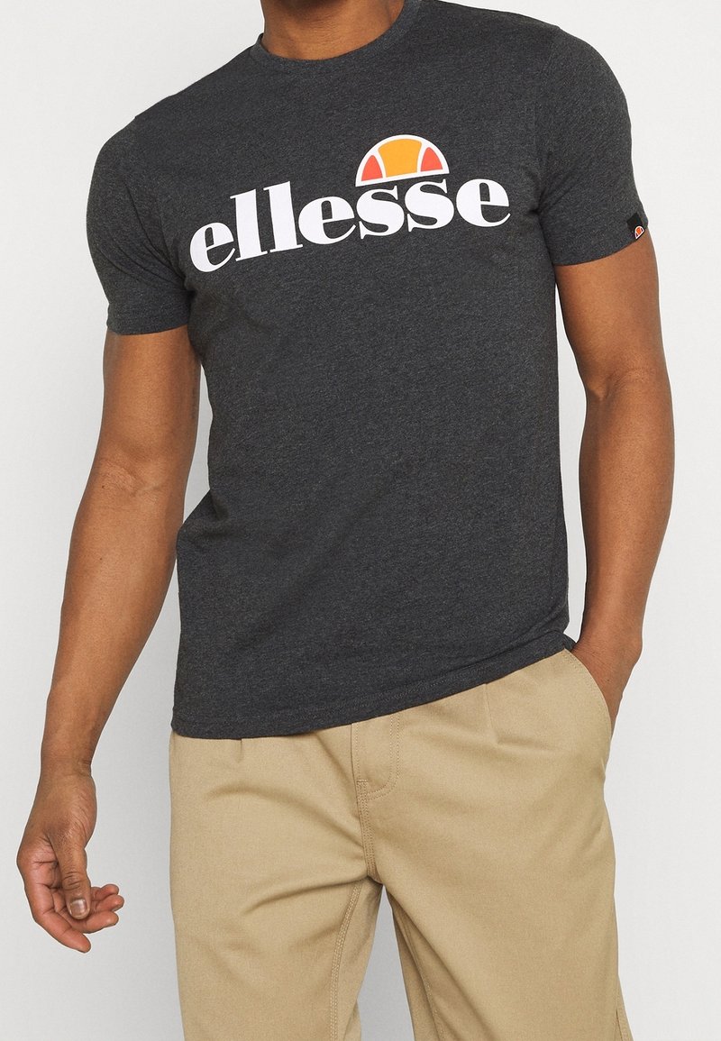 Homme portant un t-shirt ellesse gris foncé avec un logo orange, associé à un pantalon beige, debout avec une main dans la poche.