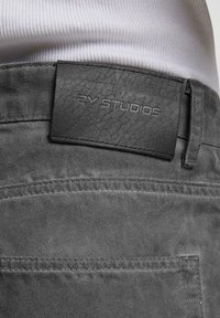 Harmaat kangashousut, joissa on tekstiilinen musta nahkalappu, johon on painettu "2Y STUDIOS". Suunnittelussa on hienovaraisia ompeluyksityiskohtia.