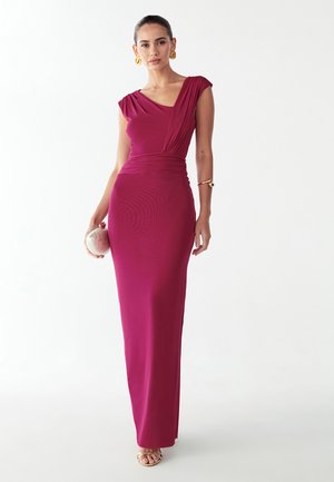 Strakke lange jurk van diep magenta stof, met een gedrapeerde halslijn, kapmouwtjes en een slanke silhouet. Gouden accessoires maken de look compleet.
