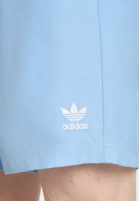 Světlé modré sportovní šortky vyrobené z hladkého materiálu, s bílým vyšitým logem Adidas a třemi pruhy nad ním.