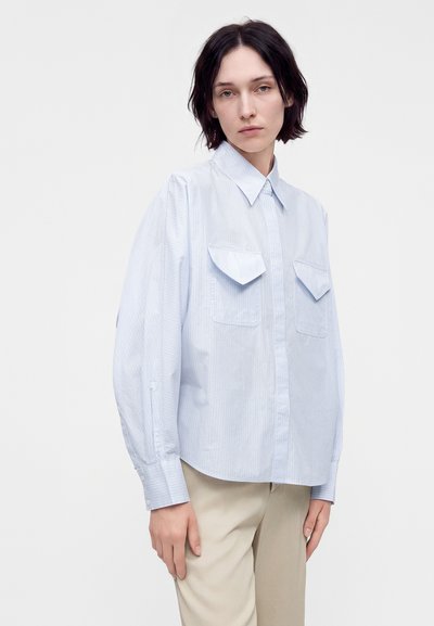 Femme aux cheveux foncés portant une chemise à rayures bleu clair avec boutons et poches à rabat et un pantalon beige sur un fond blanc uni.