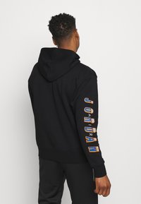 Svart hoodie med dragsko och ficka, med färgglad "JORDAN"-text längs vänstra ärmen i orange och blått. Mjuk tygstruktur.