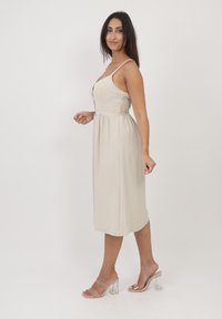 Helles beiges Midi-Kleid mit Spaghettiträgern, Knopfleiste und gerüschten Seiten. Translucente Absatzsandalen runden den Look ab.