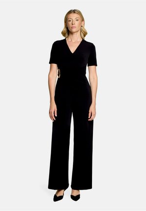 Kvinde, der står iført en sort kortærmet jumpsuit med dyb V-hals og en guldbeslagsdetalje ved taljen, kombineret med sorte spidse sko.