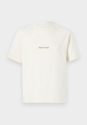 T-shirt en coton crème avec manches courtes, col rond et logo brodé "Original Studio" en doré sur la poitrine. Texture douce.