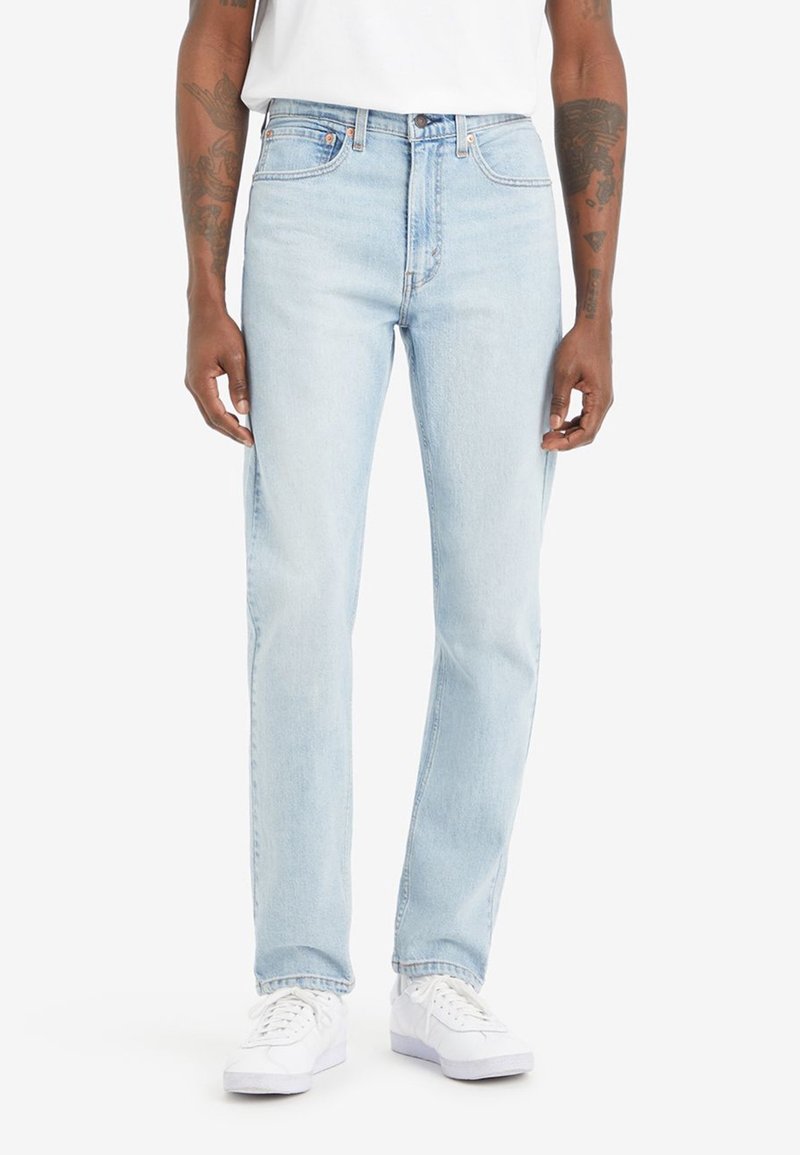 Homme portant un jean droit bleu clair et des baskets blanches, debout devant un fond blanc uni.