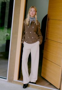 Bruine trui met witte stippen, beige wijde broek en een grijze sjaal. Staande bij een houten deur met een glazen paneel.